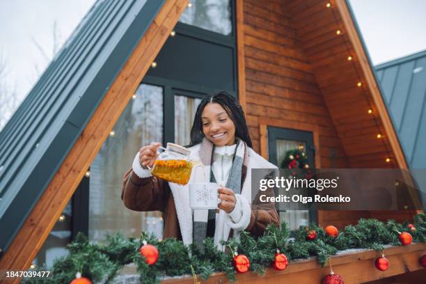 relaxed smiling woman pouring sea buckthorn tea in white mug on the porch of a decorative wooden house for christmas in glamping - white house christmas stock pictures, royalty-free photos & images