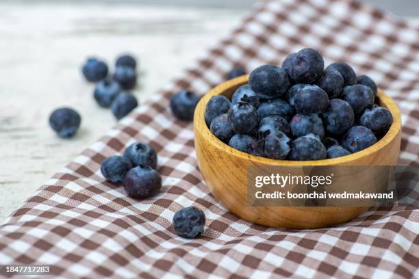 brown ceramic bowl of fresh ripe blueberries with green leaves on kitchen table - arándano fotografías e imágenes de stock