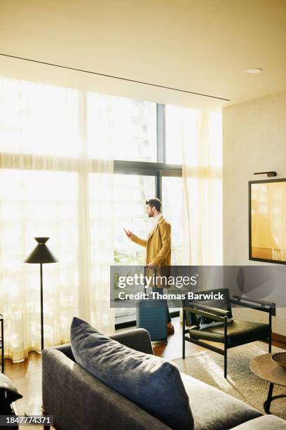 wide shot businessman checking smart phone at window of hotel suite - suitcase hotel stock pictures, royalty-free photos & images