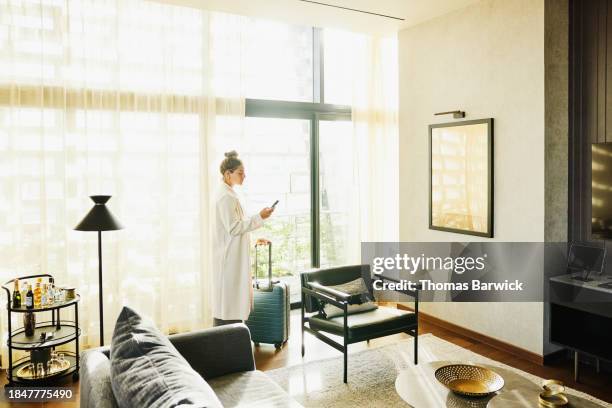 wide shot businesswoman checking smart phone at window of hotel suite - luxury hotel room stock pictures, royalty-free photos & images