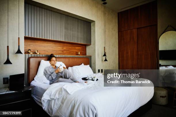 wide shot woman sitting in bed in hotel room working on smart phone - hotel stock-fotos und bilder