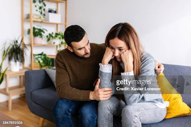 young adult man calming upset woman sitting together on sofa at home. - comfort stock pictures, royalty-free photos & images