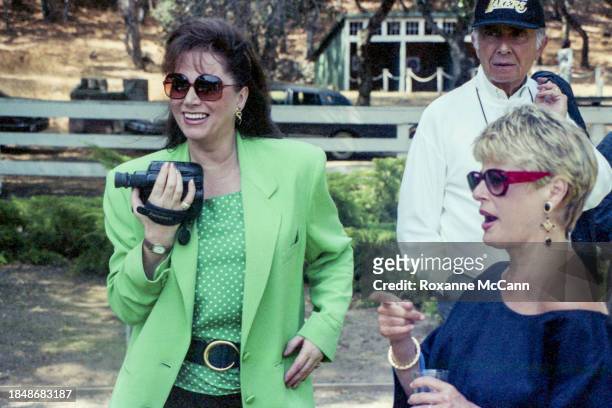 Jackie Collins, wearing sunglasses, a green suit and polka dot blouse and holding a video camera stands with Toni Howard who is wearing a blue top...