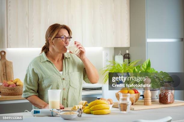 woman drinking healthy banana and protein smoothie in her kitchen - proteïnedrank stockfoto's en -beelden