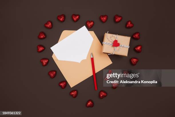 blank white paper card mockup in craft paper envelope with valentines day gift and red shiny hearts on dark chocolate brown background with copy space for text. flat lay, top view. - red card envelope stock pictures, royalty-free photos & images