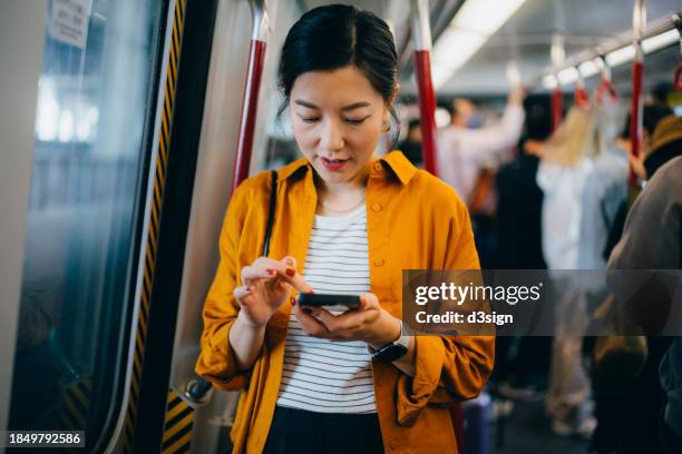 young asian businesswoman scrolling on her smartphone while riding on subway. commuting to work. travelling on public transportation in the city. lifestyle and technology - een dag uit het leven serie stockfoto's en -beelden