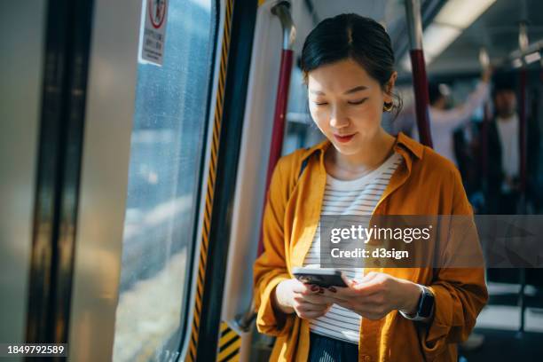 young asian woman standing by the window against sunlight in subway train while using her smartphone. travelling on public transportation in the city. lifestyle and technology - comboio de metropolitano imagens e fotografias de stock