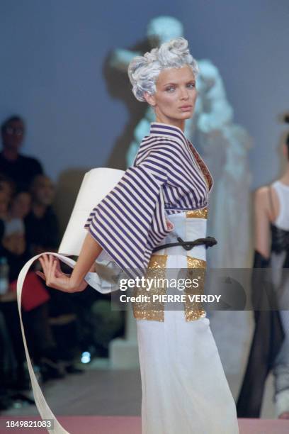 Model, with a geisha hairstyle, presents a white kimono under a sleeveless denim jacket on a long white skirt by Jean-Paul Gaultier for his...