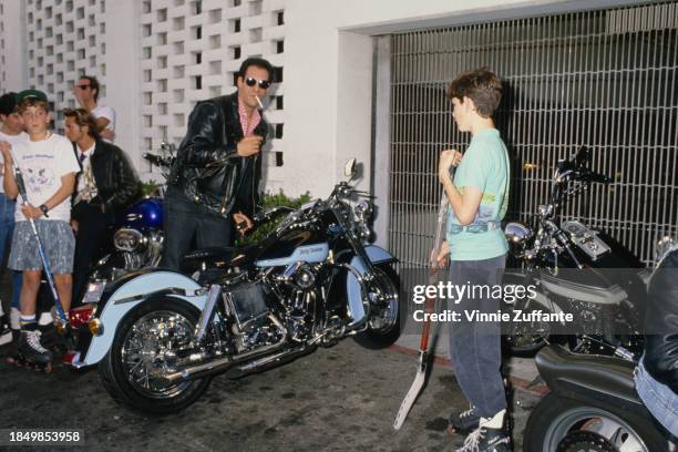 American actor and singer Robert Davi, a cigarette hanging from his mouth, wearing a black leather jacket over a red-and-white checked shirt and...