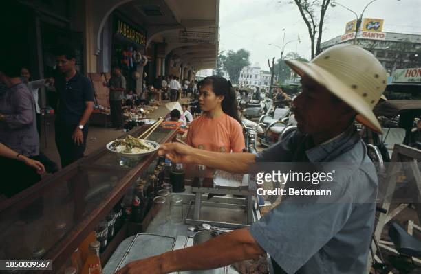 Vietnamese food vendor serving a plate of food at an open-air market in Saigon, South Vietnam, October 1967.