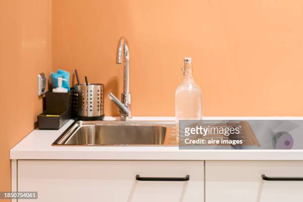 front view of a kitchen sink with a closed faucet and a glass bottle on one side, orange wall in the background. - dishwasher front photos et images de collection
