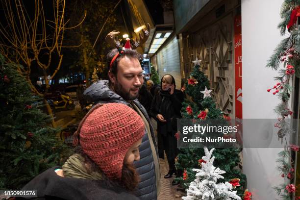 An Iranian-Christian man is wearing a Christmas accessory and looking at a Christmas tree placed on a street-side while doing Christmas shopping in...