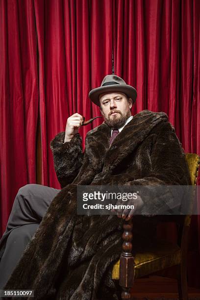 seated bearded man wearing fedora, fur coat, holding pipe - fur coat stockfoto's en -beelden