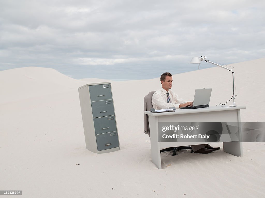 A businessman sitting on chair at desk using laptop in the middle of a desert 