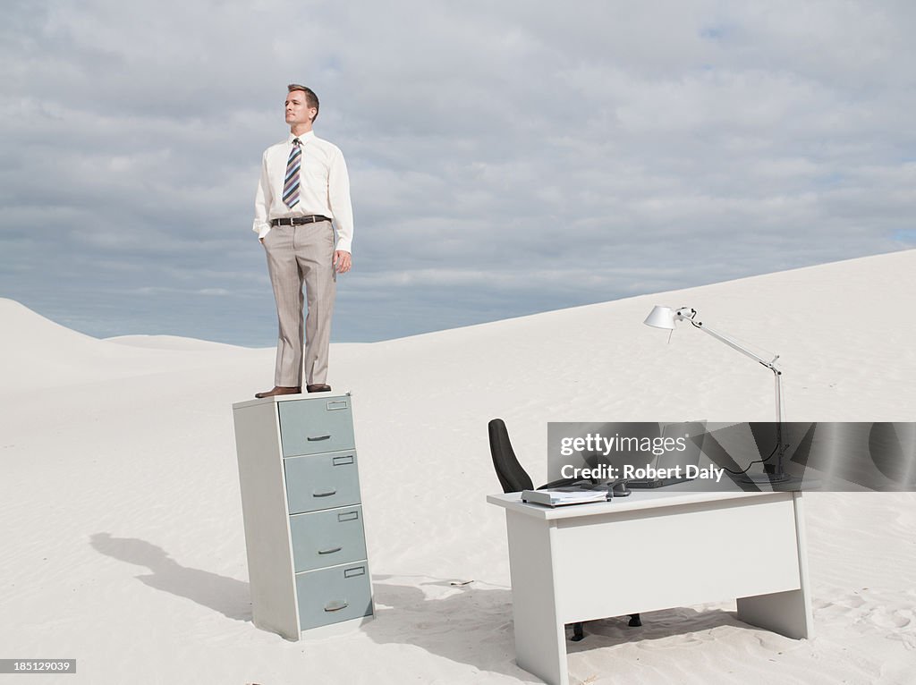 A businessman in the middle of a desert standing on his filing cabinet