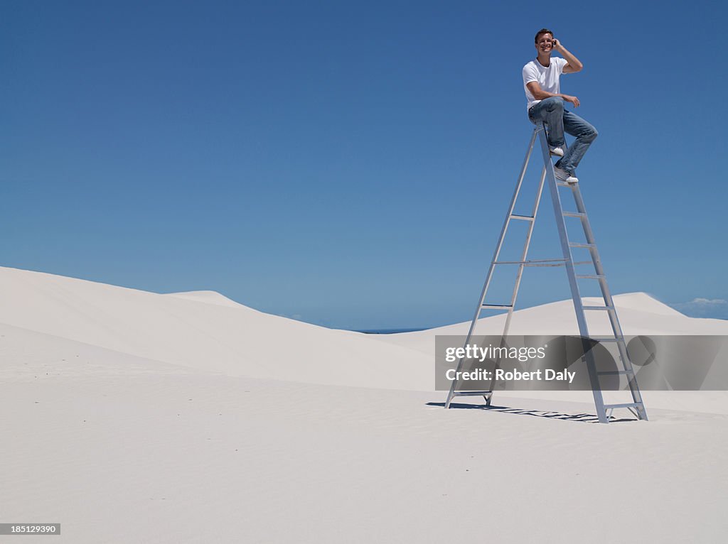 A man sitting on a ladder in the desert on a cellular phone