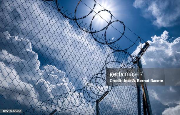 close-up of metal fence with barbed wire over a dramatic sky with the sun on the horizon. - alambrada fotografías e imágenes de stock