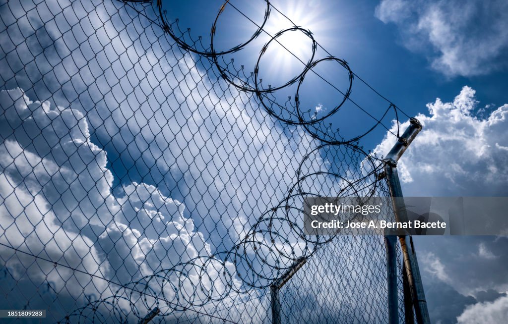 Close-up of metal fence with barbed wire over a dramatic sky with the sun on the horizon.
