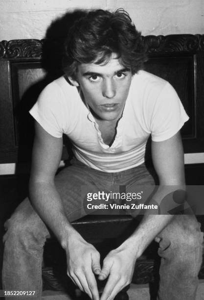 American actor Matt Dillon, wearing a white cap-sleeved t-shirt and jeans, sitting on a wooden seat with a carved backrest, United States, circa 1980.