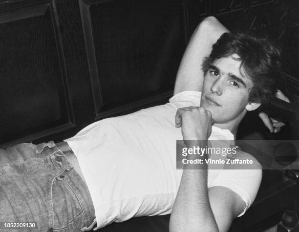 American actor Matt Dillon, wearing a white cap-sleeved t-shirt and jeans, reclining on a wooden seat, United States, circa 1983.