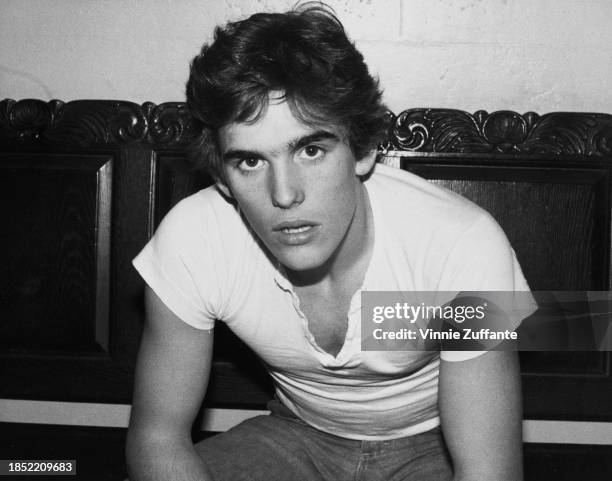 American actor Matt Dillon, wearing a white cap-sleeved t-shirt and jeans, sitting on a wooden seat with a carved backrest, United States, circa 1980.