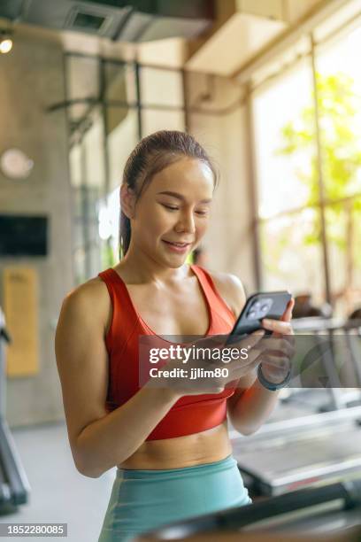 asian woman takes break from stationary bike training, using smartphone in glowing sunshine at indoor fitness gym club - peloton app stock pictures, royalty-free photos & images