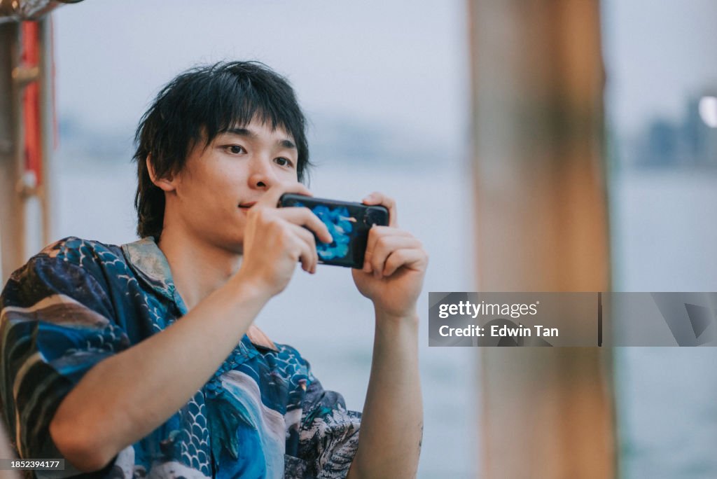 Young Asian man looking out through the window taking photos while traveling by ferry at Victoria Harbour