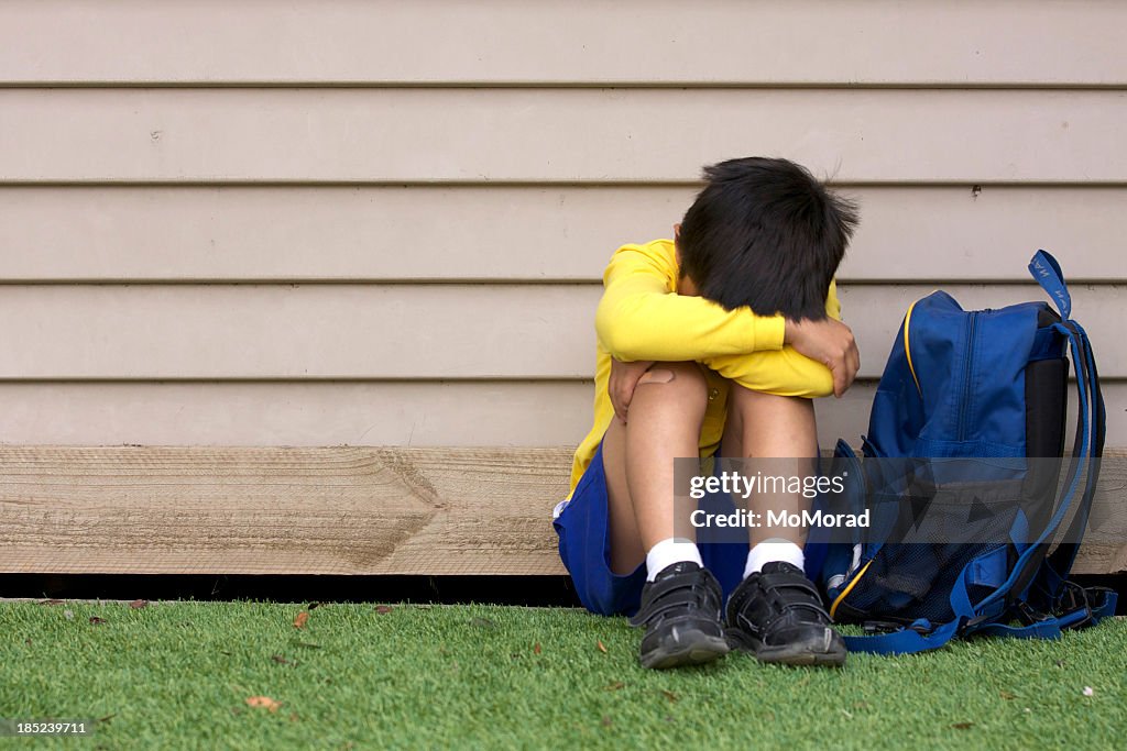 Bullied young boy sitting on grass with his head in his lap