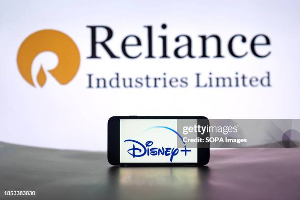 In this photo illustration, the Disney+ logo is seen displayed on a mobile phone screen with Reliance Industries Limited in the background.