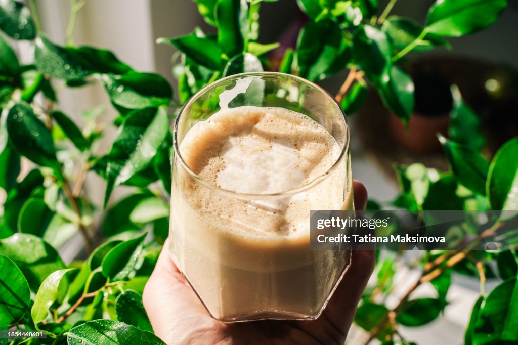 Glass of cold handmade cappuccino in hand on plants background. The little pleasures of home.