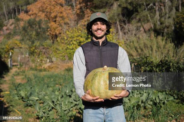 medium shot of a happy caucasian portuguese male holding a giant pumpkin in a community garden looking at the camera and smiling - giant camera stock pictures, royalty-free photos & images