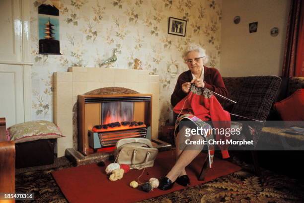 1st JUNE: View of an elderly woman knitting a Fair Isle style jumper by a gas fire in the living room of a cottage on one of the Shetland Islands in...