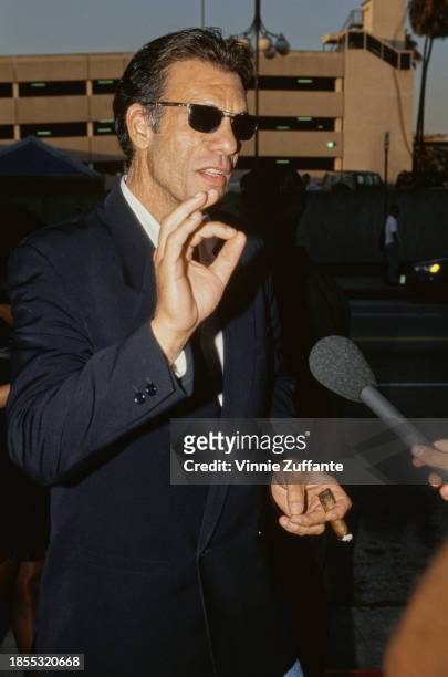 American actor and singer Robert Davi, wearing a black double-breasted blazer and sunglasses, makes the 'okay' gesture with his right hand, a lit...