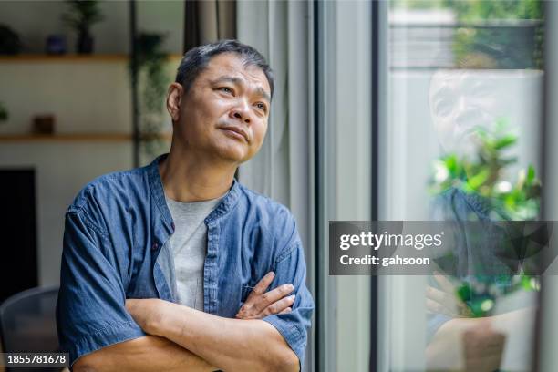 thoughtful man looking through window at home - midlife crisis stock pictures, royalty-free photos & images
