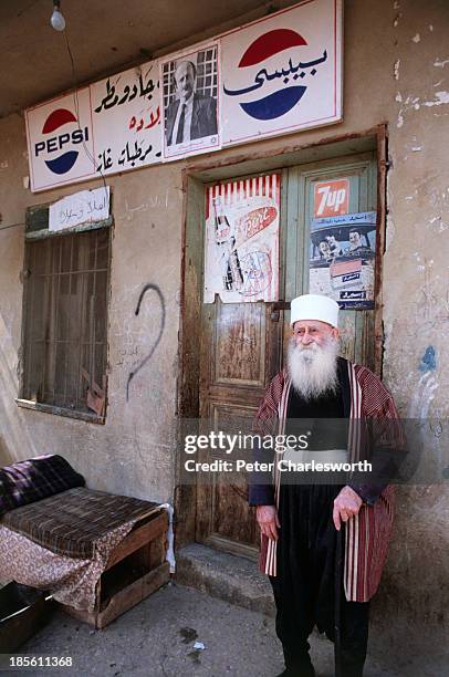 In a small village in the Chouf Mountains, a Druze Sheikh in traditional clothes stands in front of a shop front with advertising hoardings for Pepsi...