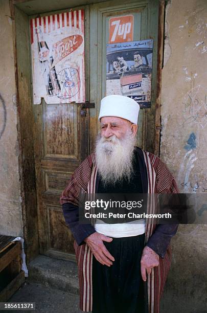In a small village in the Chouf Mountains, a Druze Sheikh in traditional clothes stands in front of a shop front with advertising hoardings for Sport...