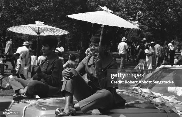 View of two women, holding parasols or umbrellas and seated on the hood of an automobile, while watching the Bud Billiken Day parade, Chicago,...