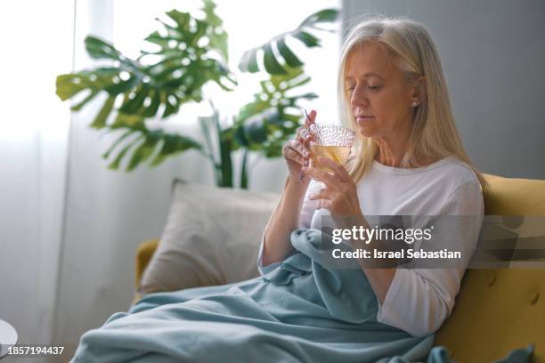 sick mature woman, sitting on the couch at her house under a blanket, drinking tea. - symptom stock-fotos und bilder