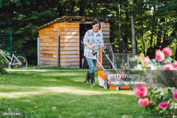 gardener yard upkeep. mature woman in work attire, using a lawnmower to cut the grass, focus on woman on background - gardening shed stock pictures, royalty-free photos & images