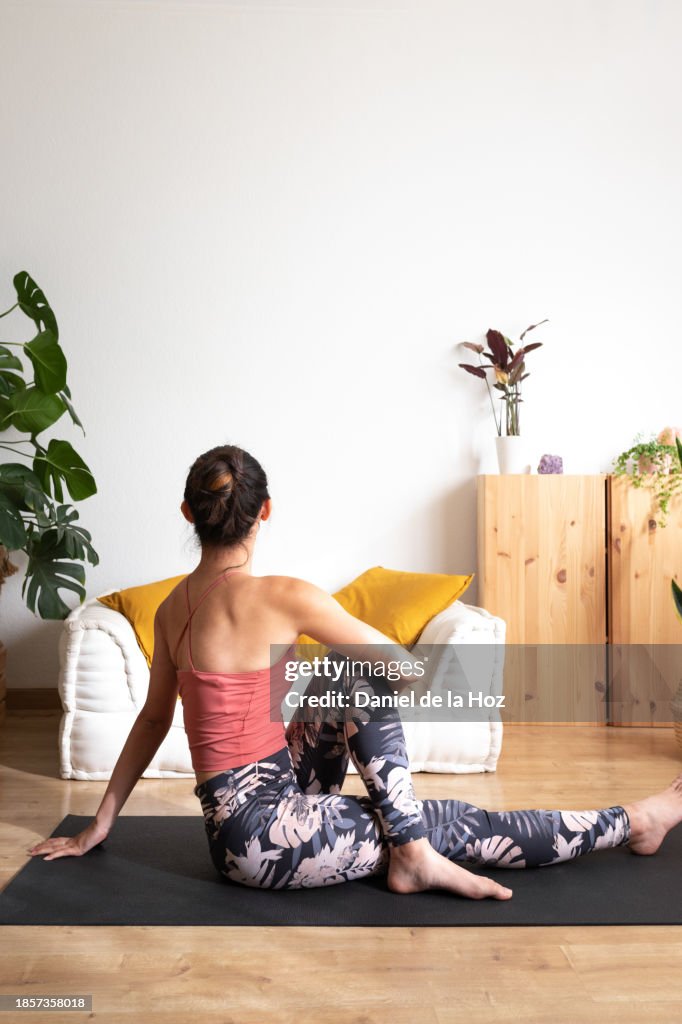 Rear view of young woman doing yoga at home. Doing seated spinal twist at home living room. Vertical image. Copy space.