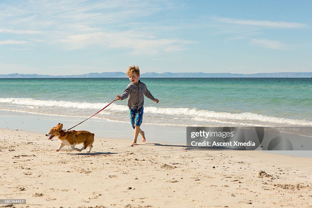 Young boy runs dog on beach, surf behind