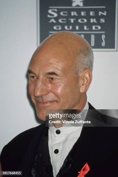 British actor Patrick Stewart, wearing a white collarless shirt with black buttons beneath a black jacket, on which is pinned an AIDS awareness...