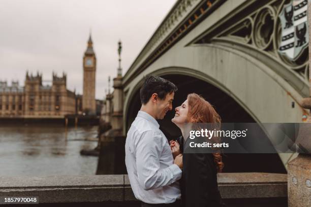 happy and young tourist couple kissing and enjoying a romantic getaway in iconic streets of london city against big ben, england, united kingdom - film romantico foto e immagini stock