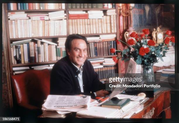 The anchorman Emilio Fede is smiling and holding a newspaper while seated at the desk of his office; behind him a bookcase. Italy, 1990.