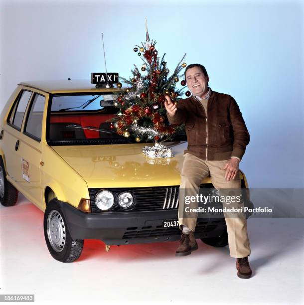Italian actor and director Alberto Sordi smiling sitting on the bonnet of a taxi beside a Christmas tree on the set of the film Il tassinaro. 1983.