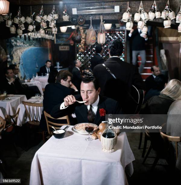 Italian actor and director Alberto Sordi eating a dish of spaghetti sitting at the table of a restaurant in the film Smoke Over London. Many flasks...