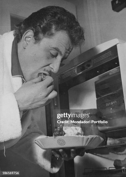 Italian singer-songwriter and actor Domenico Modugno in a bathrobe eating a dessert in front of an open fridge. 1958.