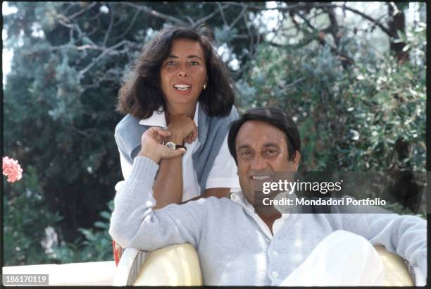 The anchorman Emilio Fede is smiling while seated on an armchair and touching a wrist of his wife, who is standing behind him. Capri , Italy, 1982.