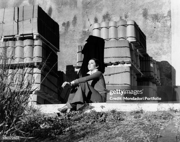 The Italian actress and singer Daniela Goggi is smiling while seated down with her arms on her knees on a sunny day in front of piles of building...