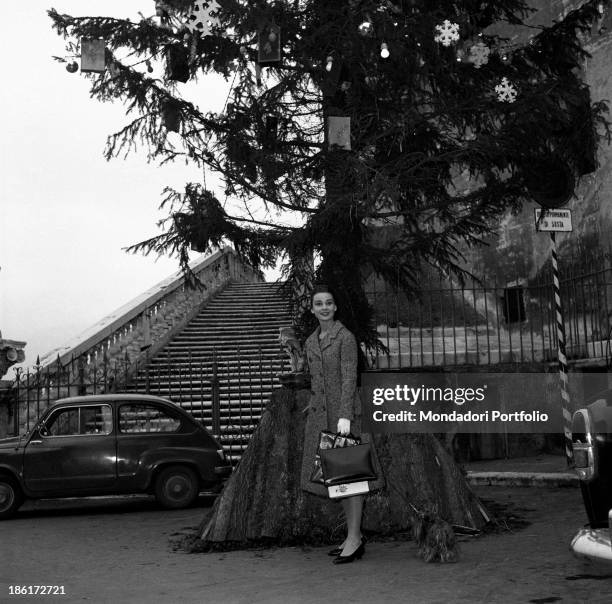 British actress Audrey Hepburn standing in front of a big tree decorated for Christmas. Behind her, a stately staircase. Rome, 1950s.